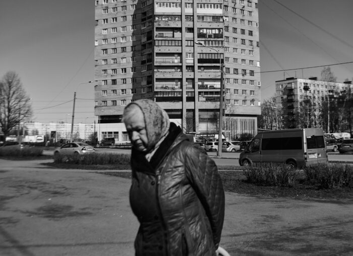 Schwarzweißaufnahme einer alten Frau mit dicker Jacke vor einem Plattenbau