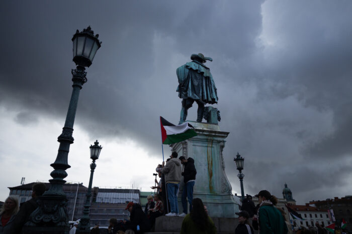 Statur eines Mannes in neuzeitlicher Kleidung auf einem Platz. An seinem Sockel Personengruppe mit Palästinafahne, links davon Straßenlaternen im Hintergrund Gebäude und grauer Himmel